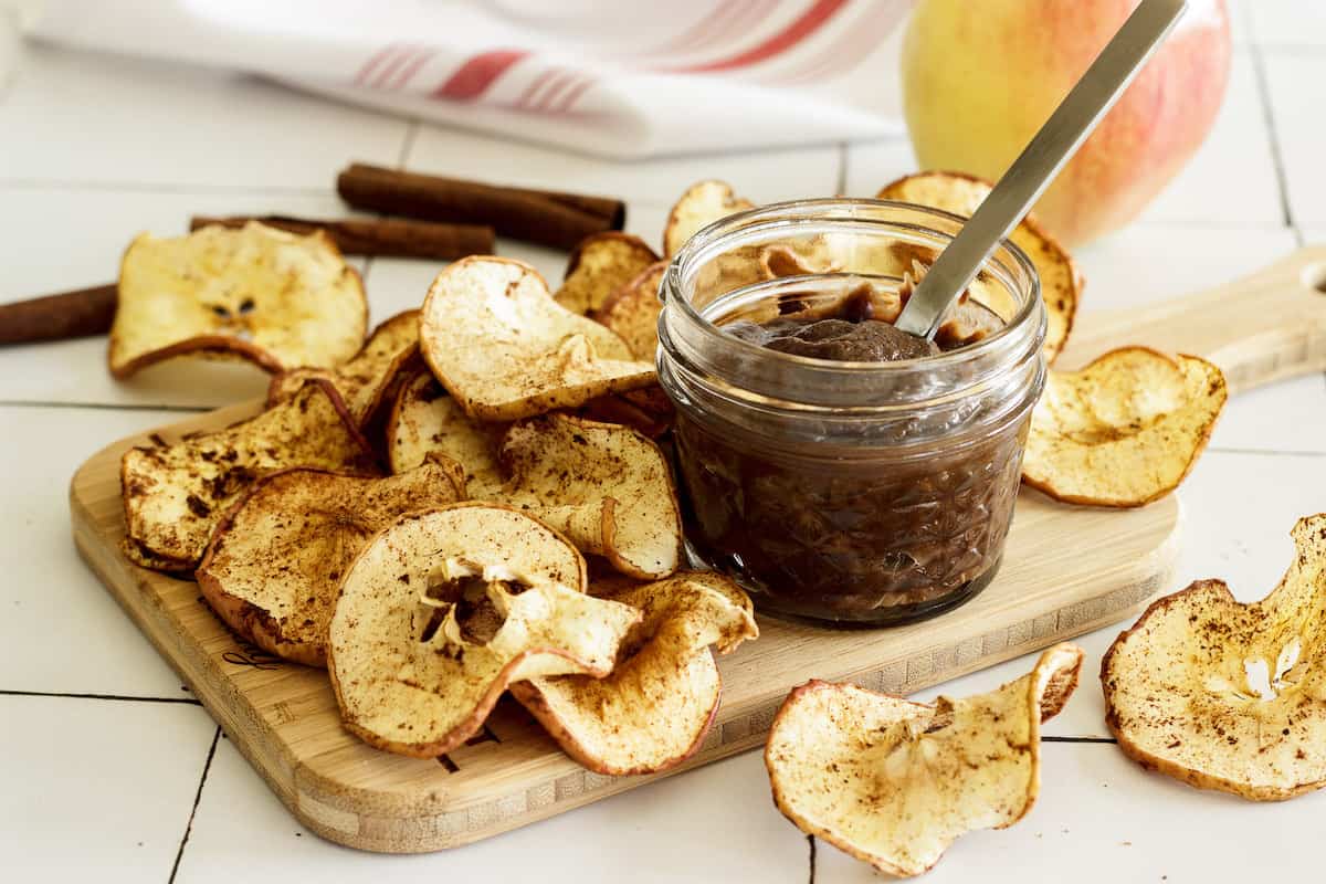 A board with a pile of apple chips and a small jar of chocolate dip.