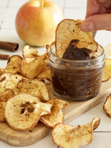 A board with a pile of air fryer apple chips and a small jar of chocolate dip. A hand is dipping an apple chip into the chocolate dip. An apple and cinnamon stick in the background