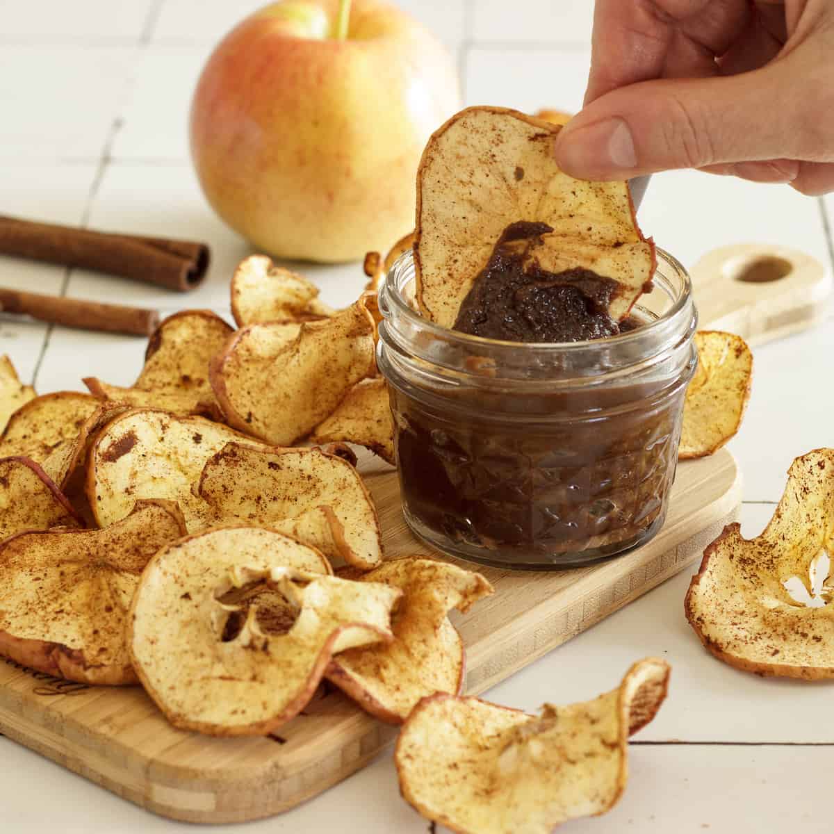 A board with a pile of air fryer apple chips and a small jar of chocolate dip. A hand is dipping an apple chip into the chocolate dip. An apple and cinnamon stick in the background