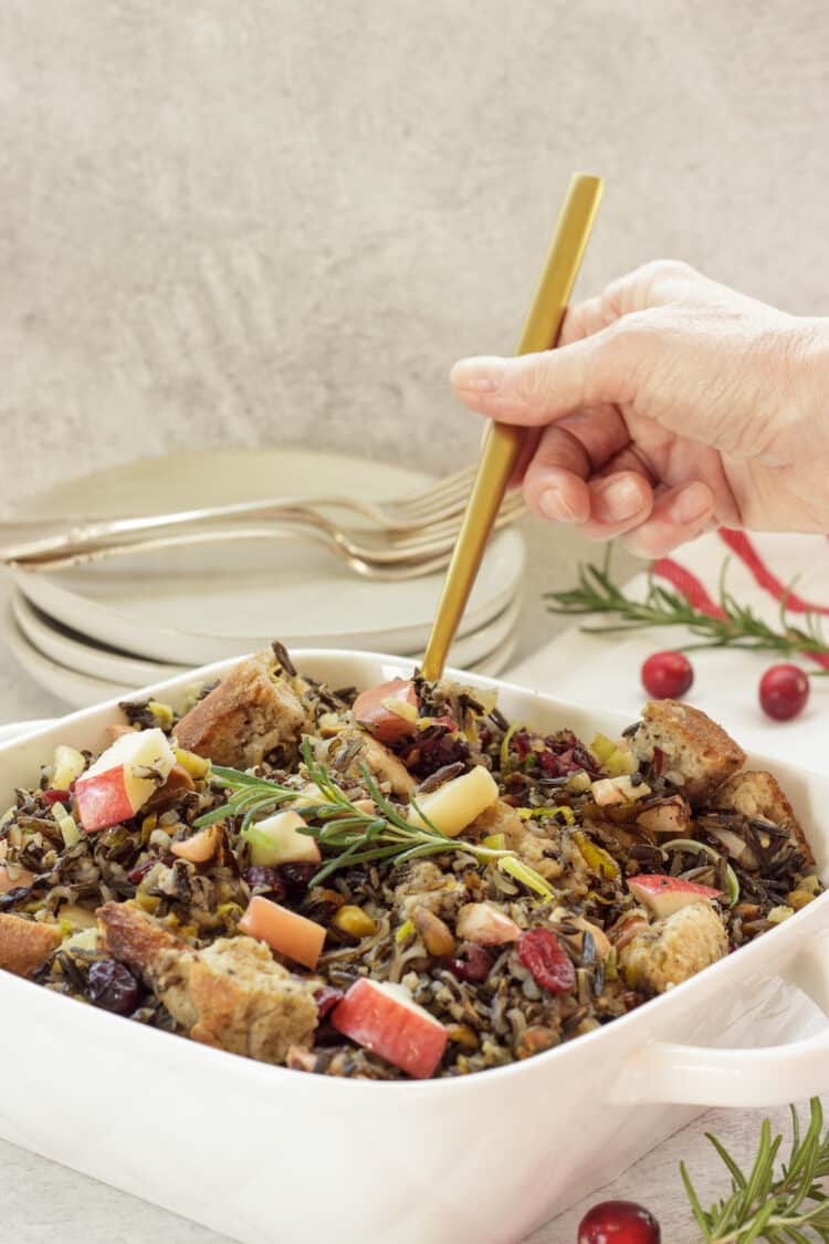 A person holding a gold spoon scooping wild rice stuffing casserole from a white casserole dish