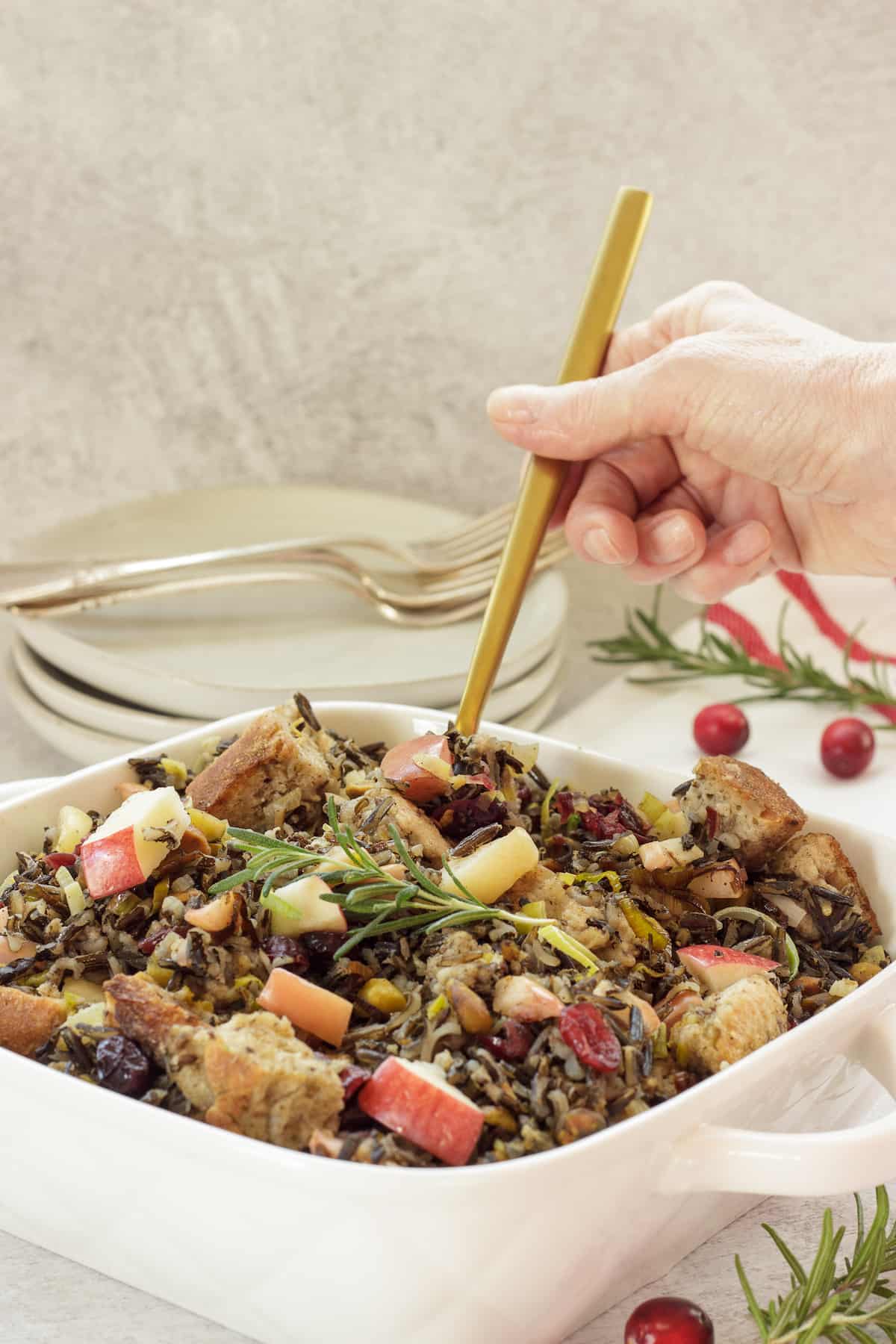 A person holding a gold spoon scooping wild rice stuffing casserole from a white casserole dish