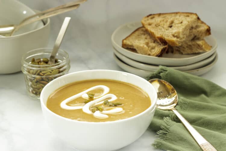 A white bowl of roasted acorn squash soup garnished with pepitas and a swirl of sour cream. A stack of white plates with bread in the background.