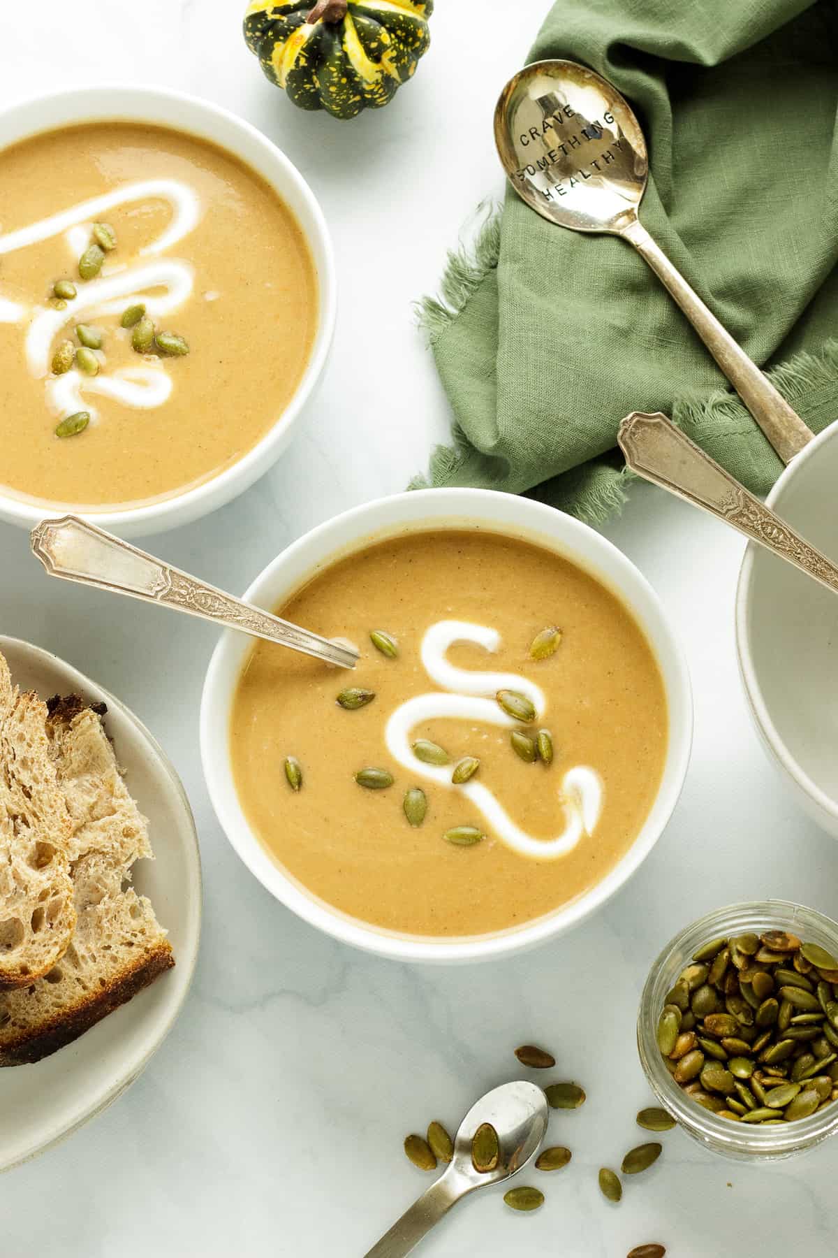 White bowls with roasted acorn squash soup topped with pumpkin seeds and a swirl of yogurt. Sliced bread and a container of pumpkin seeds are in the background.