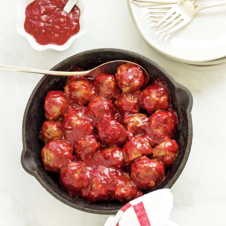 A cast iron pan full of mini meatballs covered with cranberry sauce. A bowl of sauce and a stack of white plates are next to the pan.