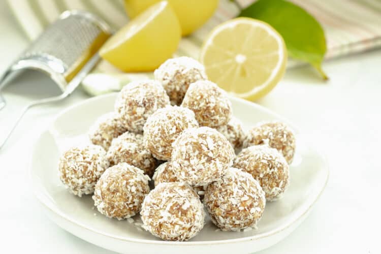 A plate of coconut covered energy balls on a white plate with cut lemons in the background
