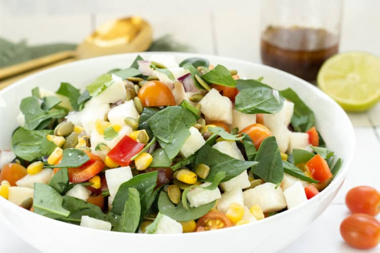 A white bowl of jicama salad. A gold serving spoon, jar of dressing, cut lime, and grape tomatoes are in the background.