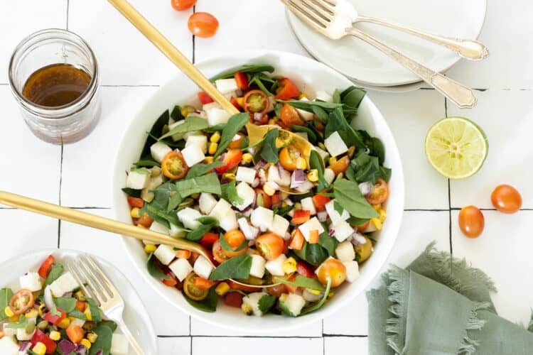 A white bowl of jicama salad with two serving spoons. A jar of dressing, cut lime, grape tomatoes, and small plate of salad surround the main bowl.