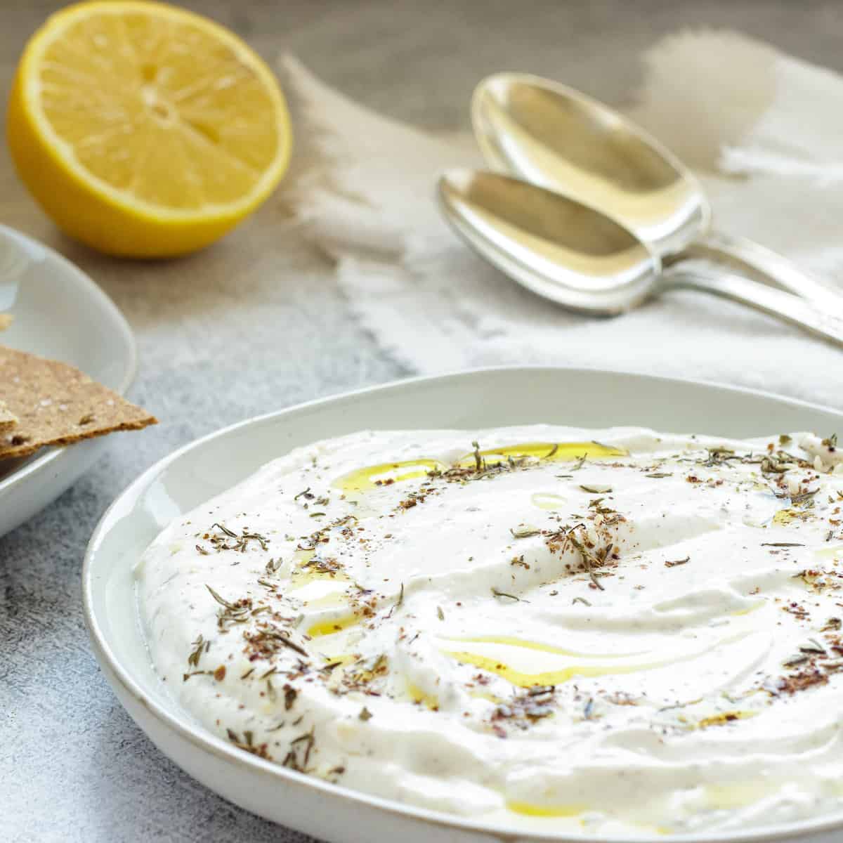 A white serving plate with whipped feta dip. A cut lemon, spoons, and plate of crackers are in the background.