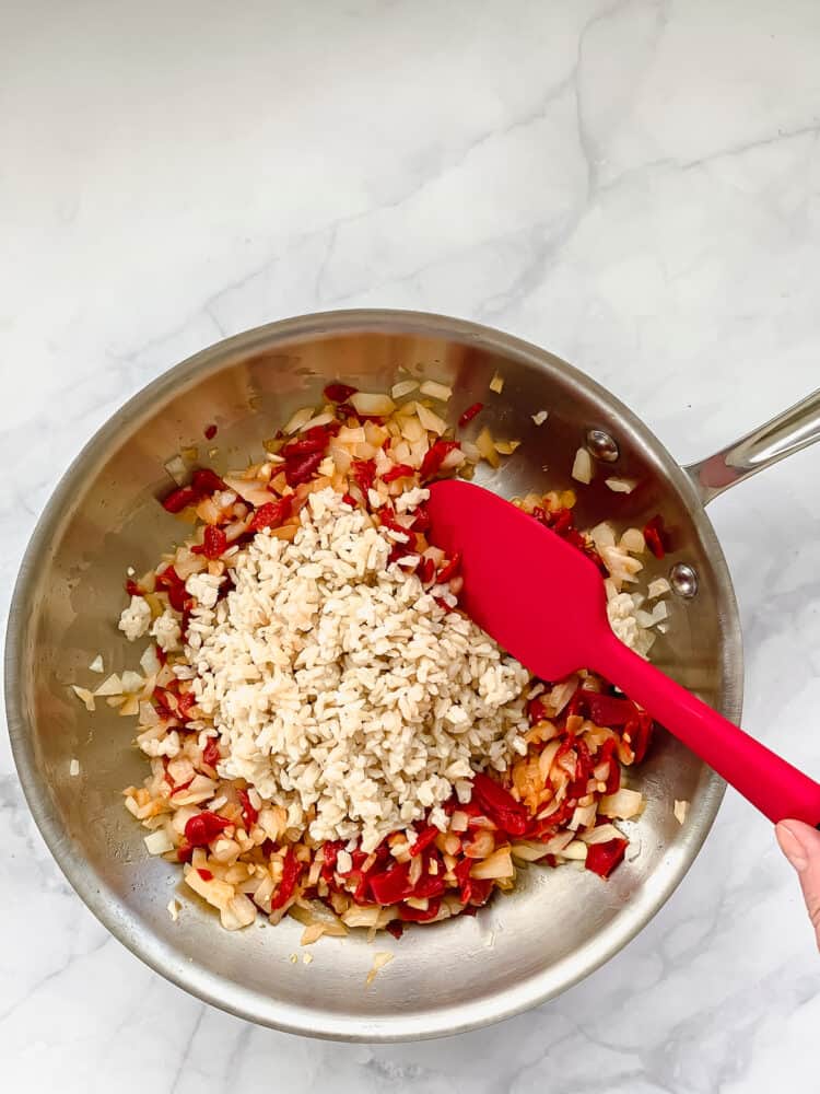 A frying pan with chopped red peppers, onions, and rice. A red spatula stirring everything.