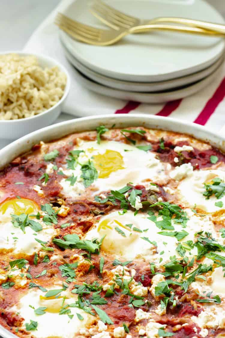 A pan of shakshuka (tomato and eggs) with parsley. A small bowl of rice and a stack of white plates in the background.