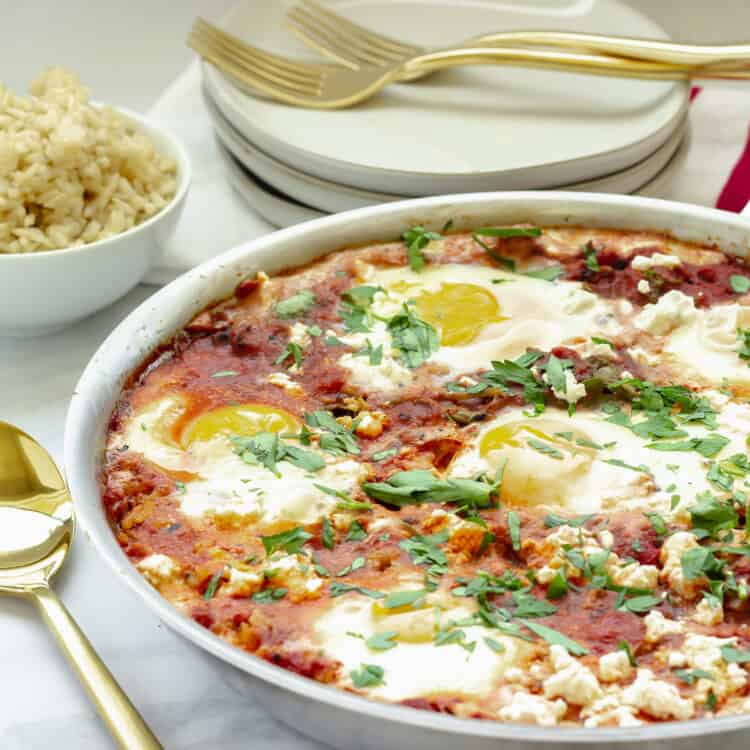 A pan of Shakshuka (tomatoes and eggs with parsley sprinkled over). A stack of white plates and gold forks and a small white bowl of rice in the background