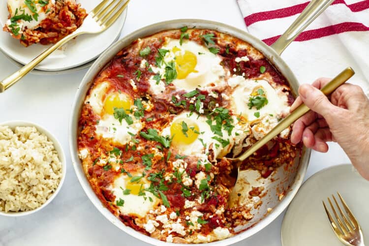 A person holding a serving spoon in a pan of Shakshuka (tomatoes and eggs). Plates, forks, and a small bowl of rice surround the pan.