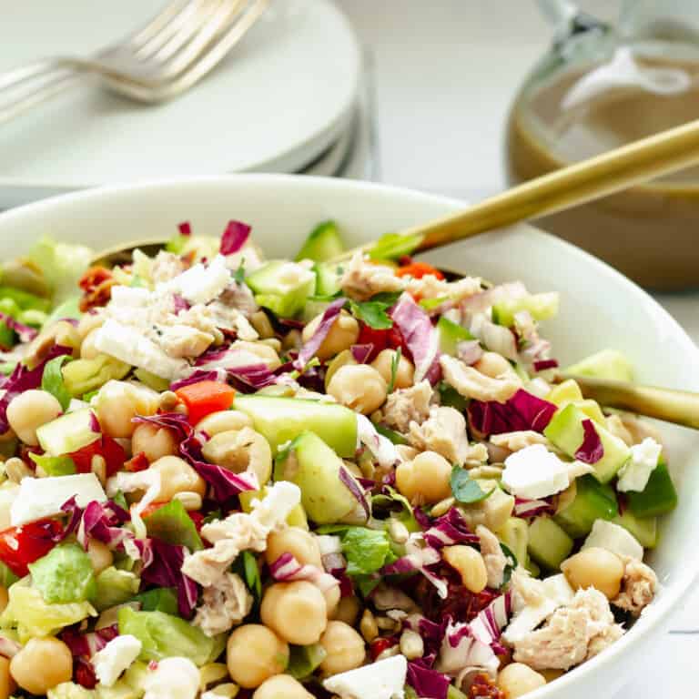 A white bowl of Mediterranean chopped salad. Serving plates, forks, and a bottle of dressing in the background.