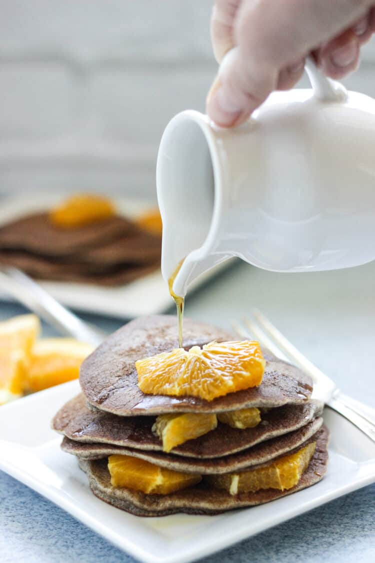 A small white pitcher with syrup being poured over buckwheat pancakes with orange segments.