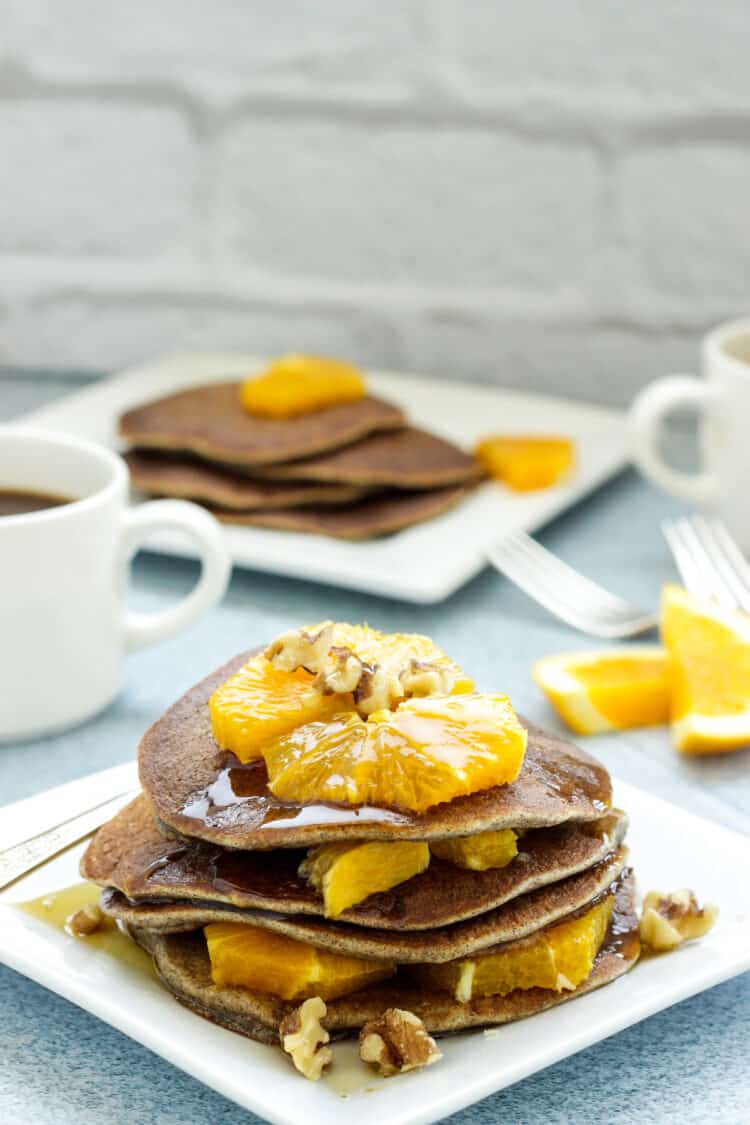 A closeup shot of white plate of buckwheat pancakes topped with orange segments. White coffee cups and a plate of pancakes in the background.