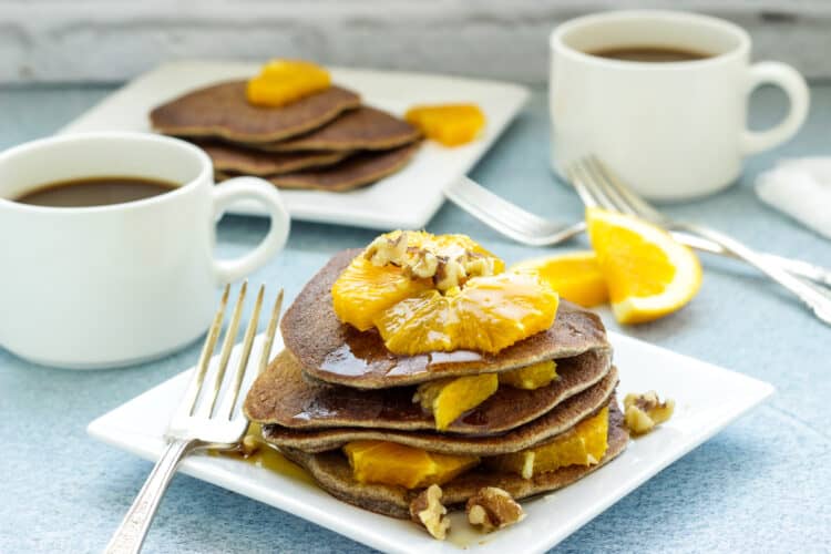 A white plate of buckwheat pancakes topped with orange segments. White coffee cups in the background.