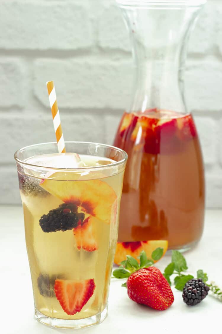 A closeup shot of a glass of iced sun tea with fruit and a pitcher of tea in the background.