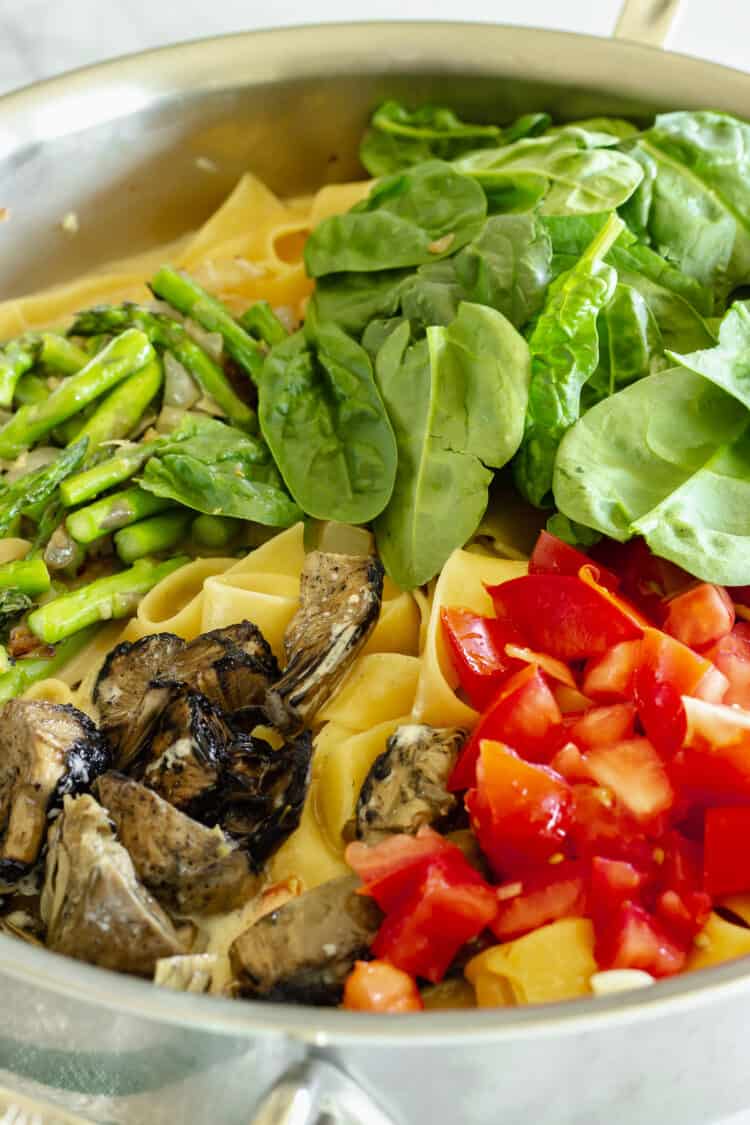 Closeup of a pan with pasta, asparagus, spinach, tomatoes, and roasted artichoke hearts.