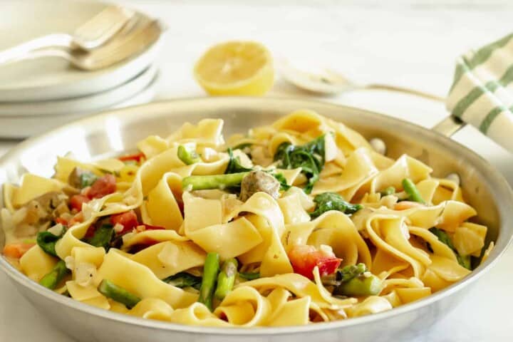 A plate of pasta with wide noodles is mixed with asparagus, spinach, tomatoes, and capers. A lemon half is in the background, along with stacked plates and cutlery on a cloth.