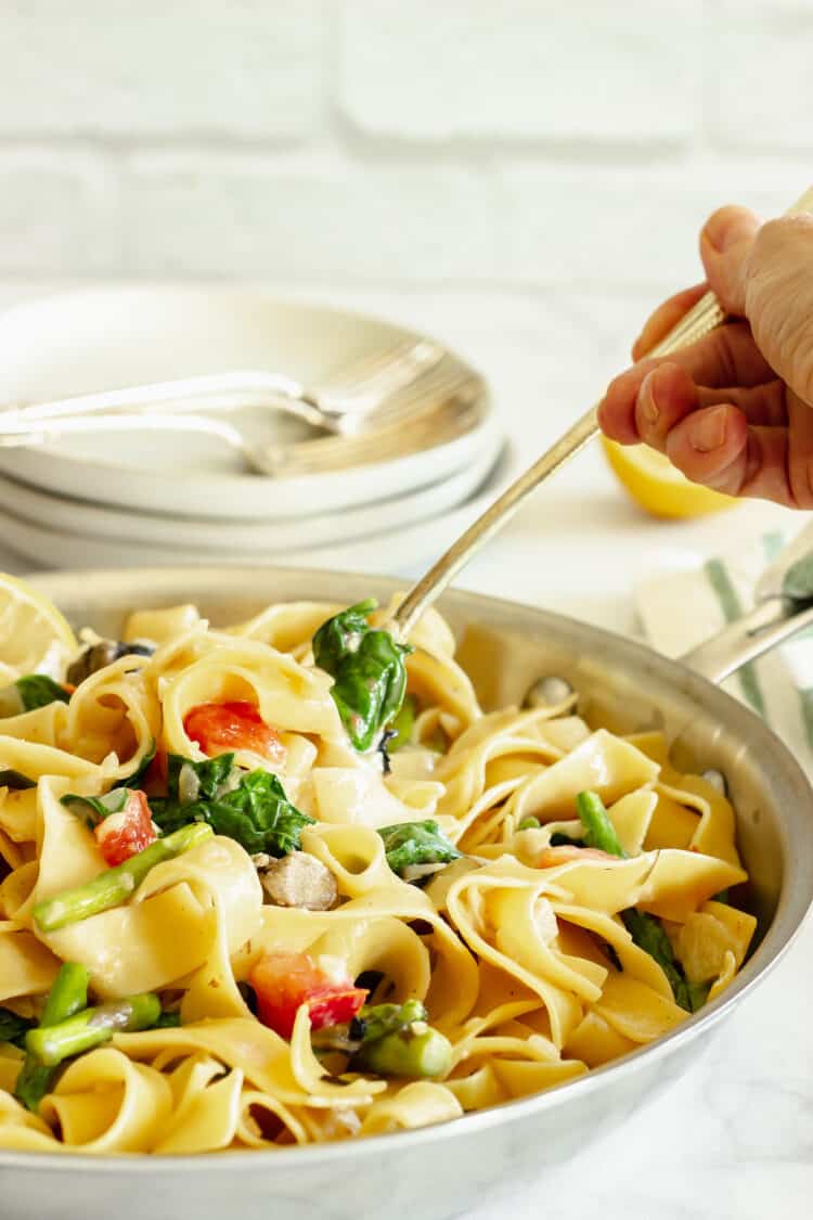 A closeup of a spoon serving pappardelle pasta primavera out of a pan.