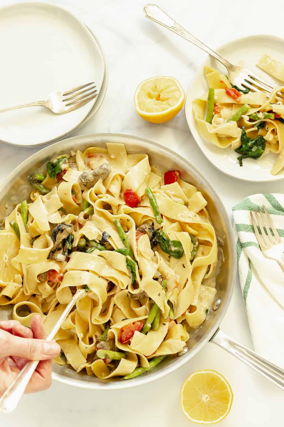 an overhead shot of a hand holding a serving spoon in a pan of pappardelle pasta primavera