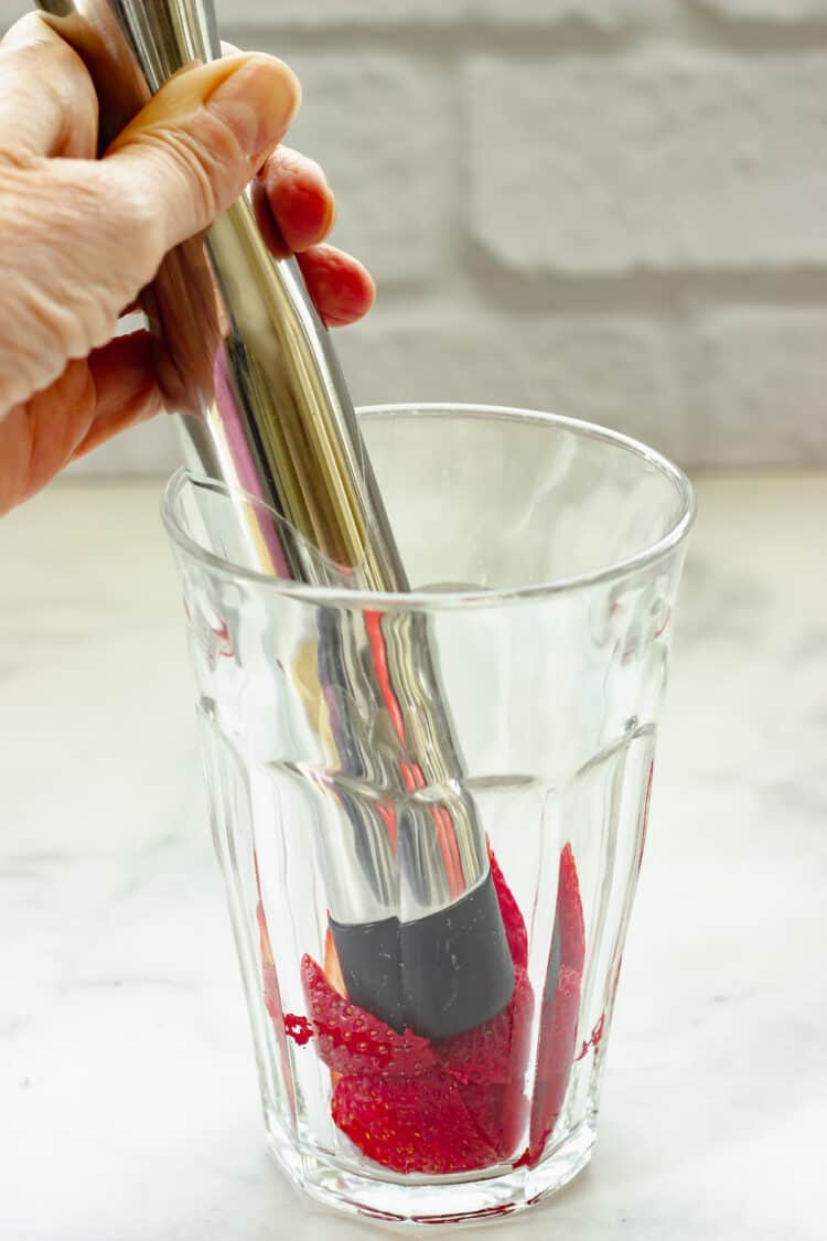 A person using a muddler to mash strawberries into a drinking glass.