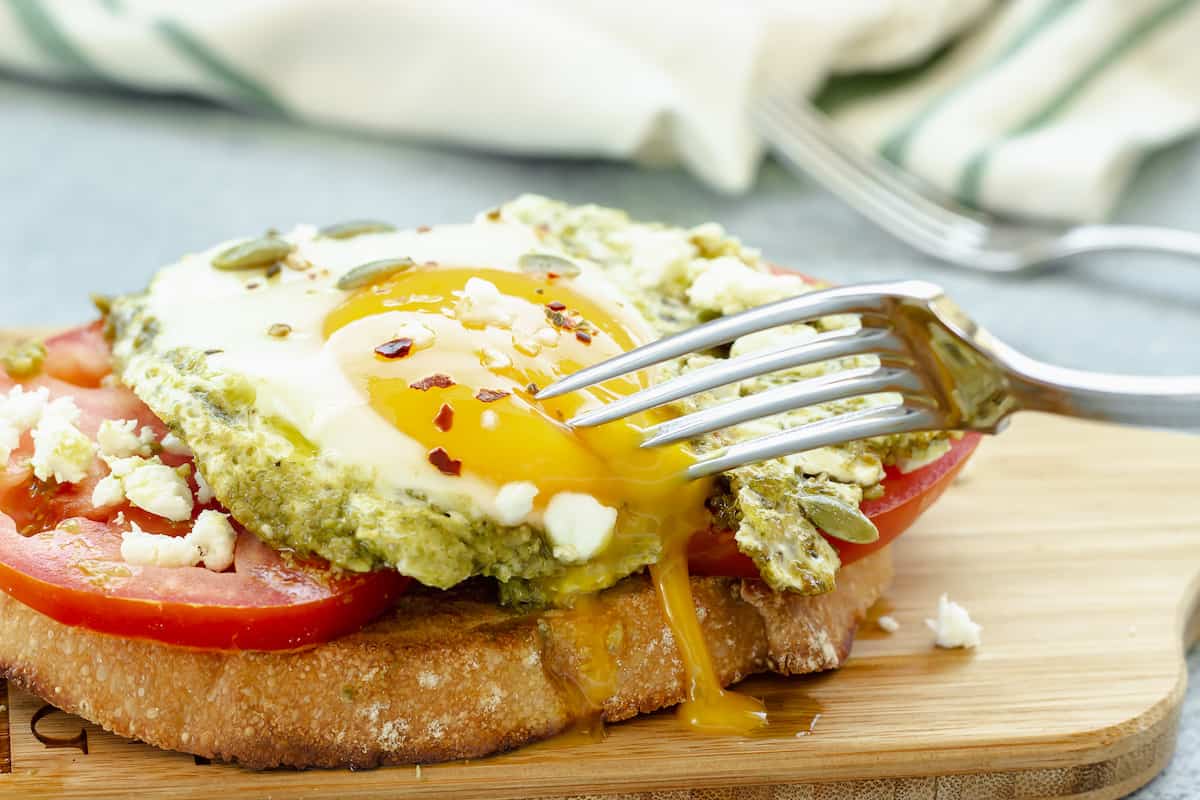 A closeup shot of pesto eggs on top of tomato and toast. A fork is cutting into a soft, drippy, egg yolk.