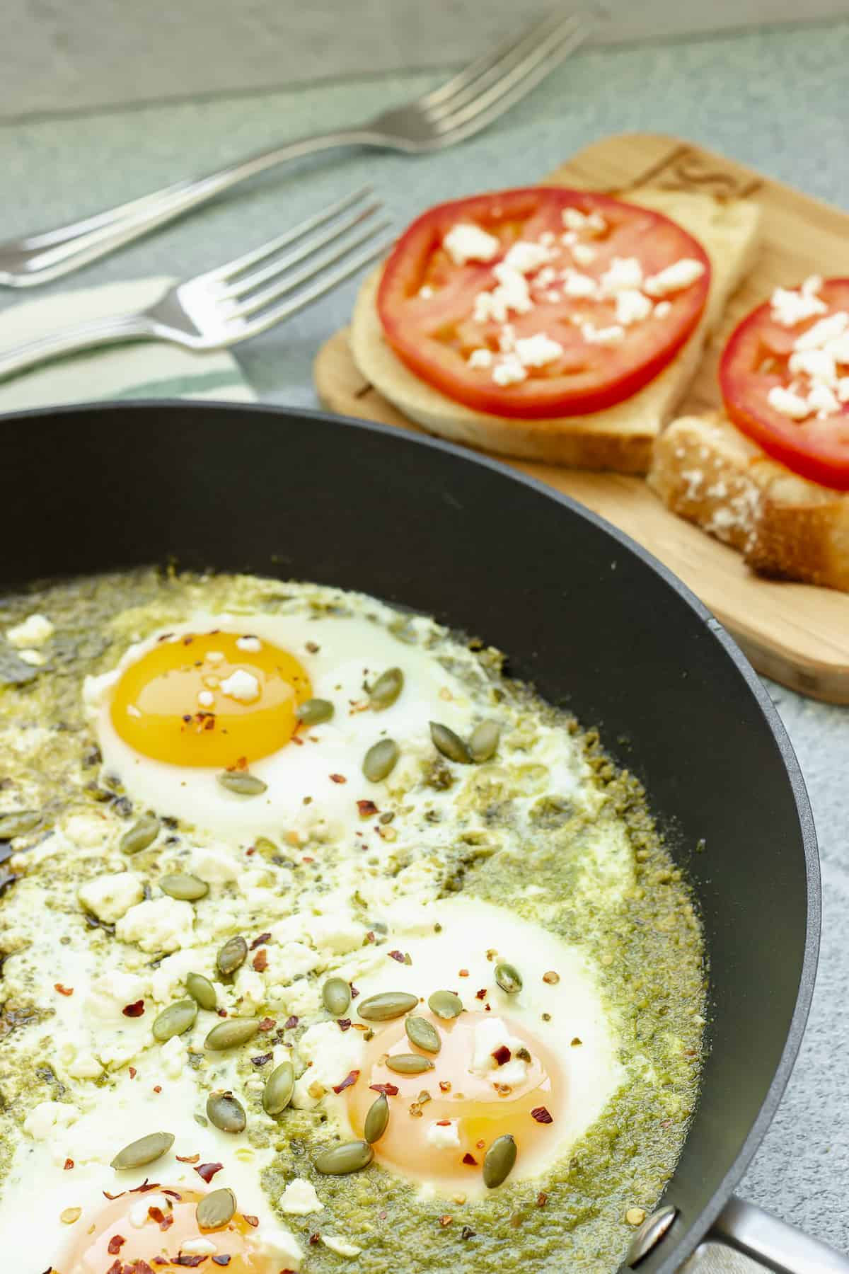 An overhead shot of a pan of pesto eggs with toast and tomato slices in the background.