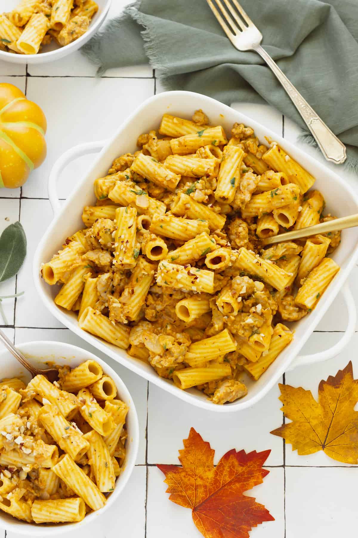 A white casserole dish and two white bowls of rigatoni with creamy pumpkin pasta sauce. 