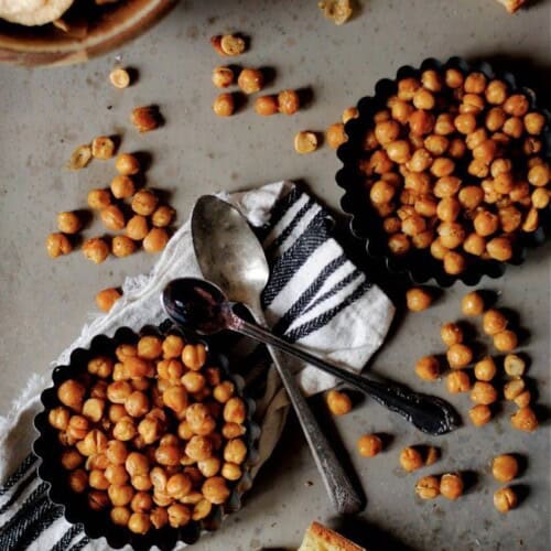 two bowls of roasted chickpeas on a tan background with a napkin and spoons.