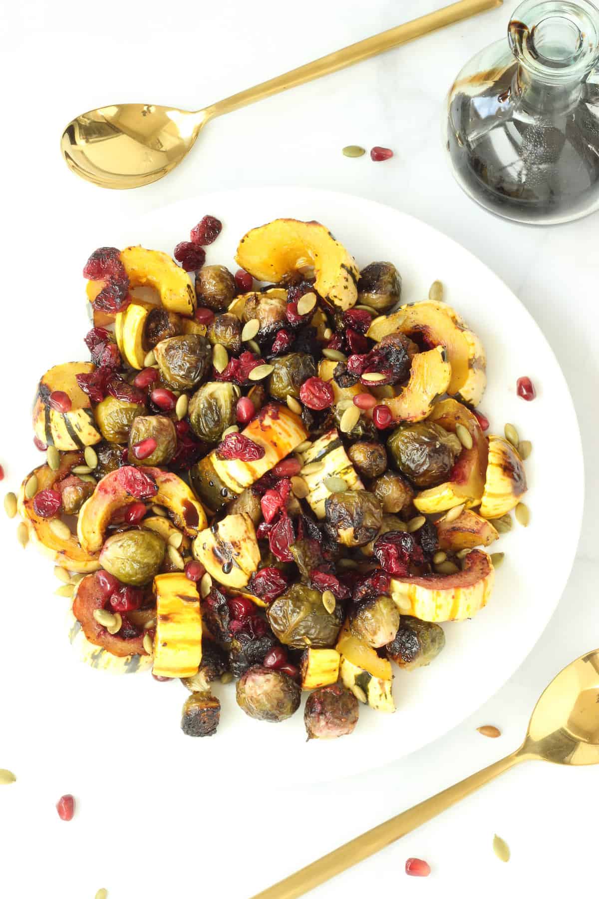 An overhead shot of roasted fall vegetables (Brussels sprouts, delicata squash, and cranberries)on a white plate with gold serving spoons on either side of the plate.