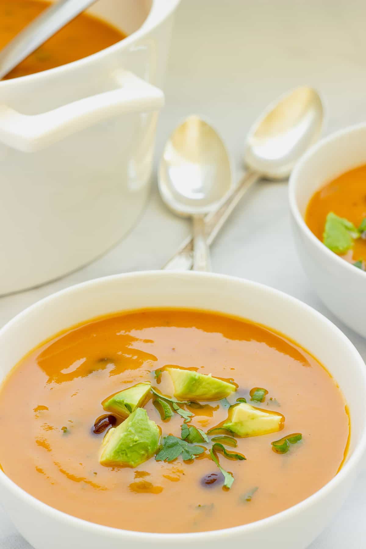 A white bowl of orange colored creamy sweet potato black bean soup garnished with diced avocado and cilantro. Spoons, a white pot of soup and a second bowl of soup are in the background. 