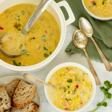 A white soup pot with chicken corn chowder. A plate of bread, two bowls of soup, spoons, and a green napkin are in the background.