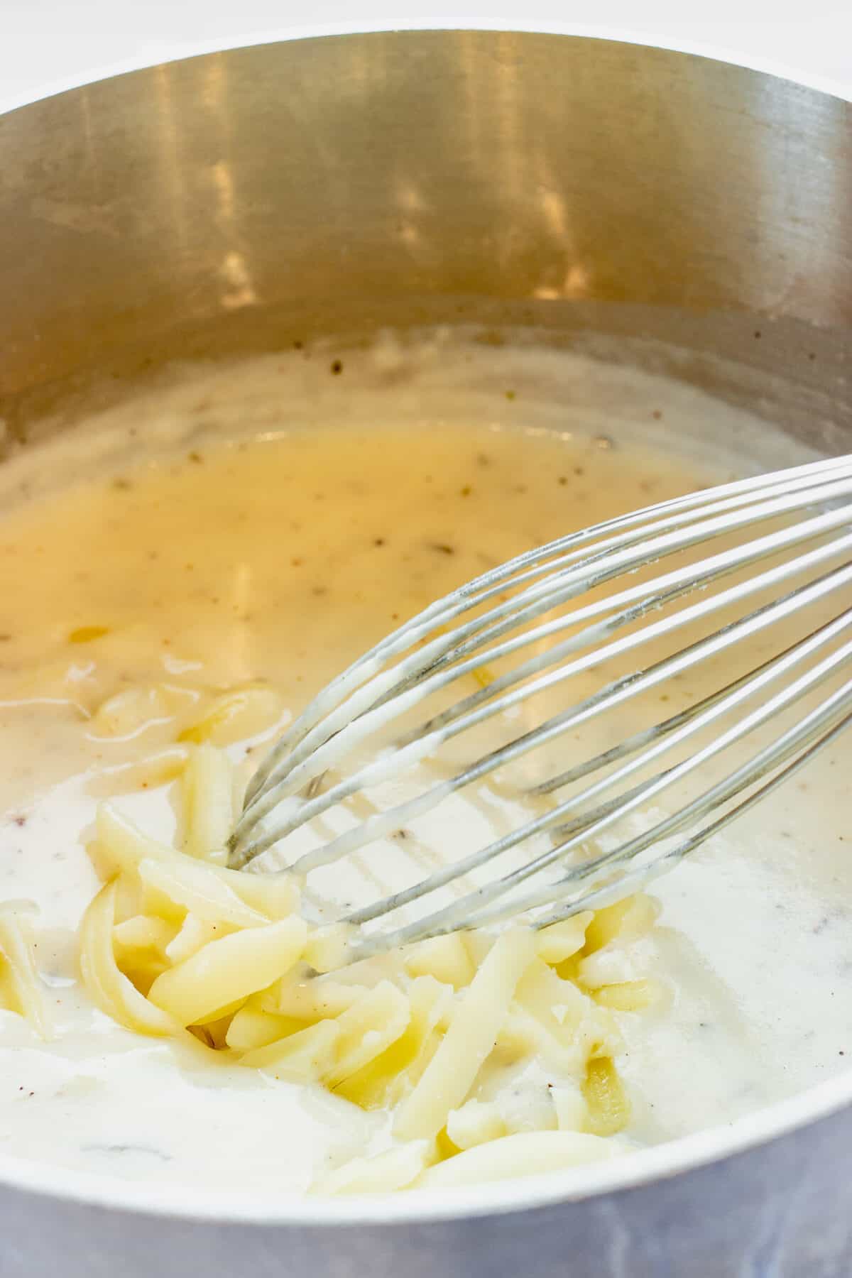 A whisk stirring shredded Gruyere cheese into a pot of Mornay sauce. Step 2 of how to make mixed vegetable casserole.