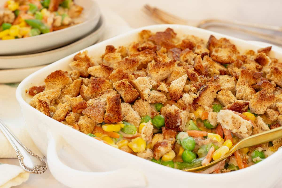 A white casserole dish with mixed vegetable casserole and a serving spoon. A stack of plates and silverware is in the background.