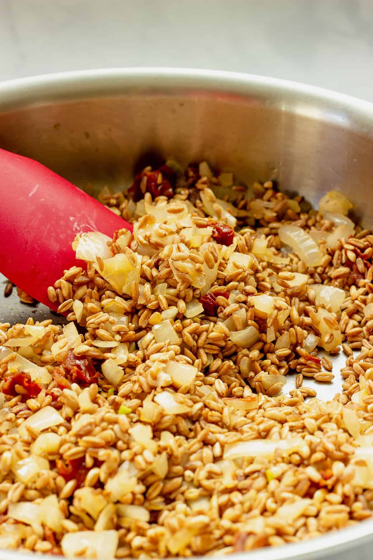 A saute pan and red spatula stirring a mixture of farro, onions, garlic, and sun-dried tomatoes.