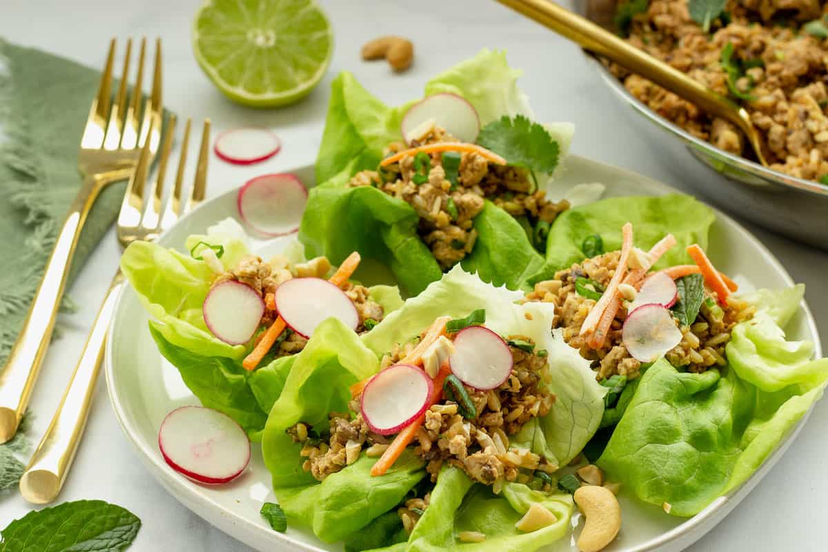 A closeup shot of a white plate with tofu lettuce wraps topped with fresh radishes, carrots, herbs, and cashews.