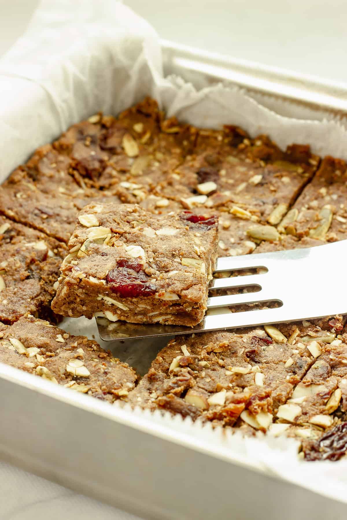 A pan of fruit and nut bars. A spatula is lifting one bar out of the pan.