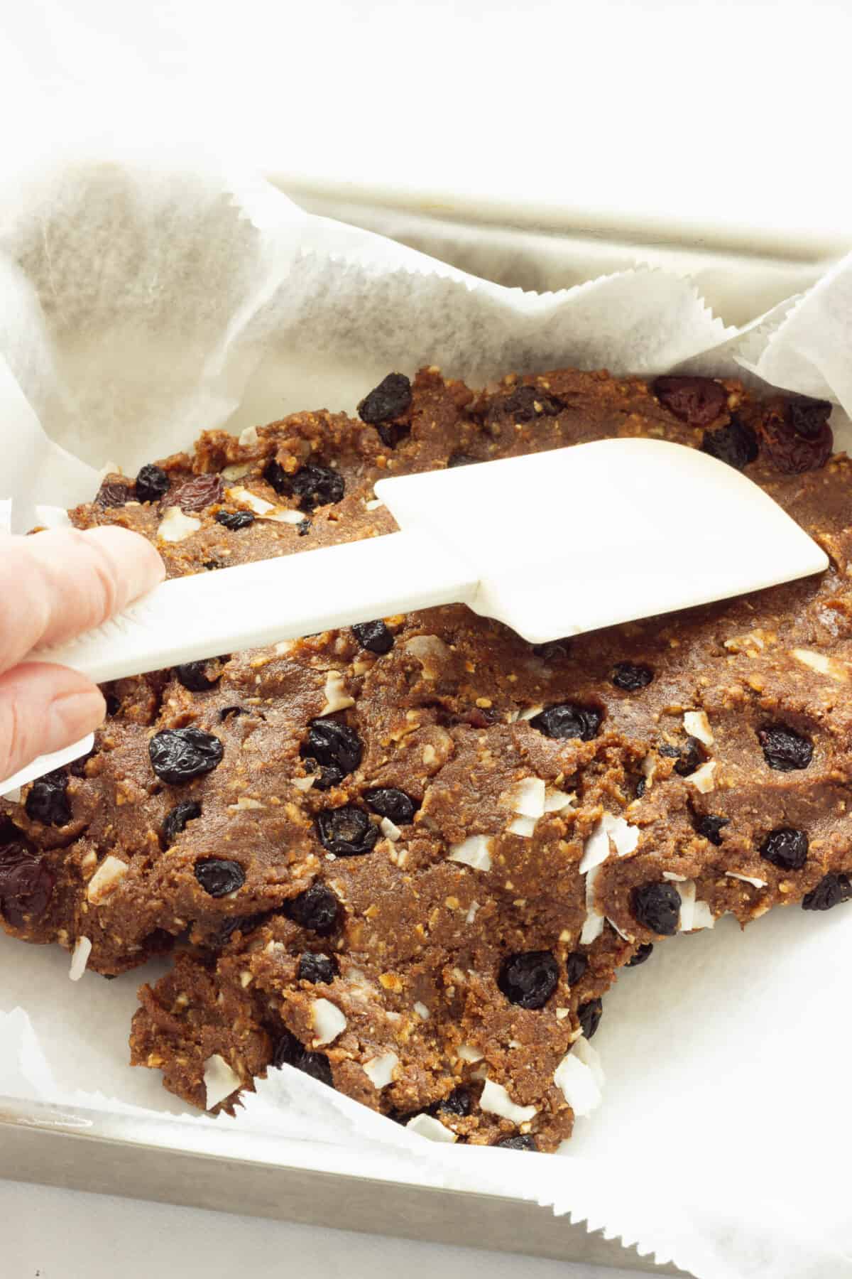 A person holding a white spatula spreading fruit and nut bar dough into a baking pan.