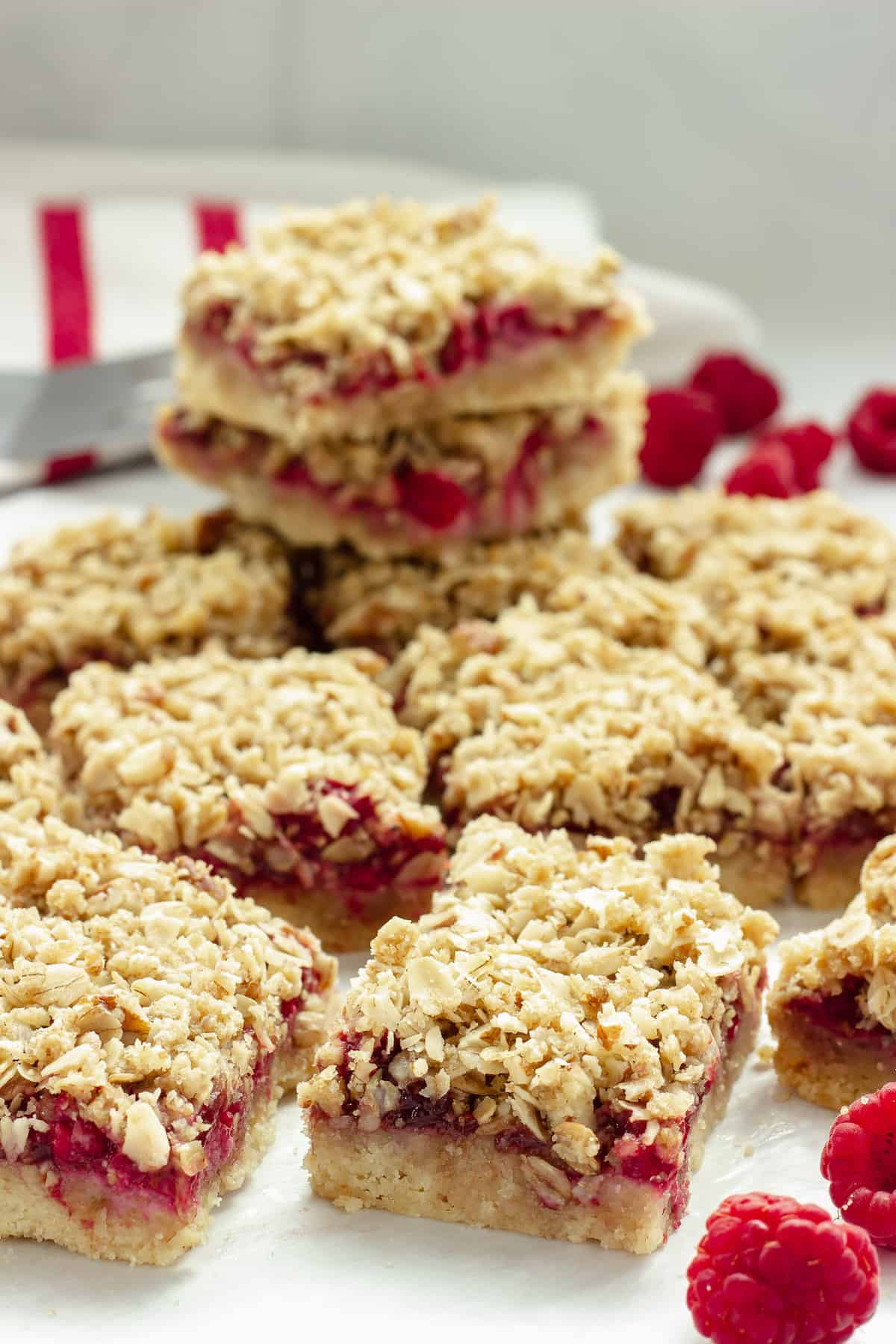 Raspberry oatmeal bars lined up on a sheet of parchment paper with fresh raspberries in the foreground and background.