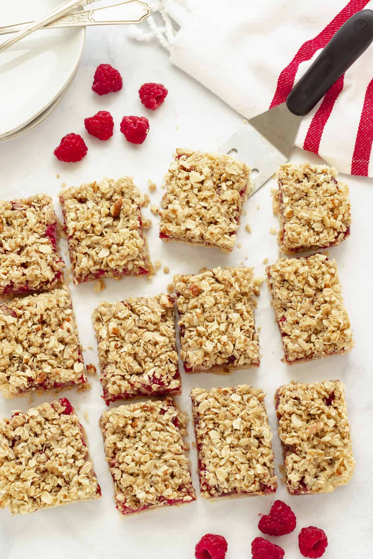 raspberry oatmeal bars cut into 12 squares. A spatula is scooping up one bar. Fresh raspberries, white plate and a red and white stripe napkin in the background.