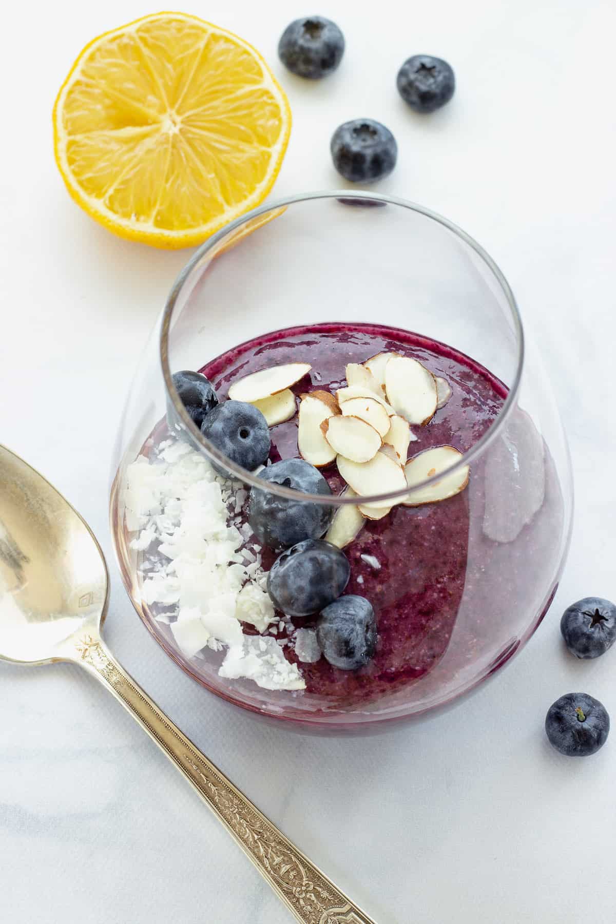 A glass of blueberry chia pudding topped with sliced almonds, blueberries, and coconut. Some fresh blueberries, a lemon, and a spoon are in the background.