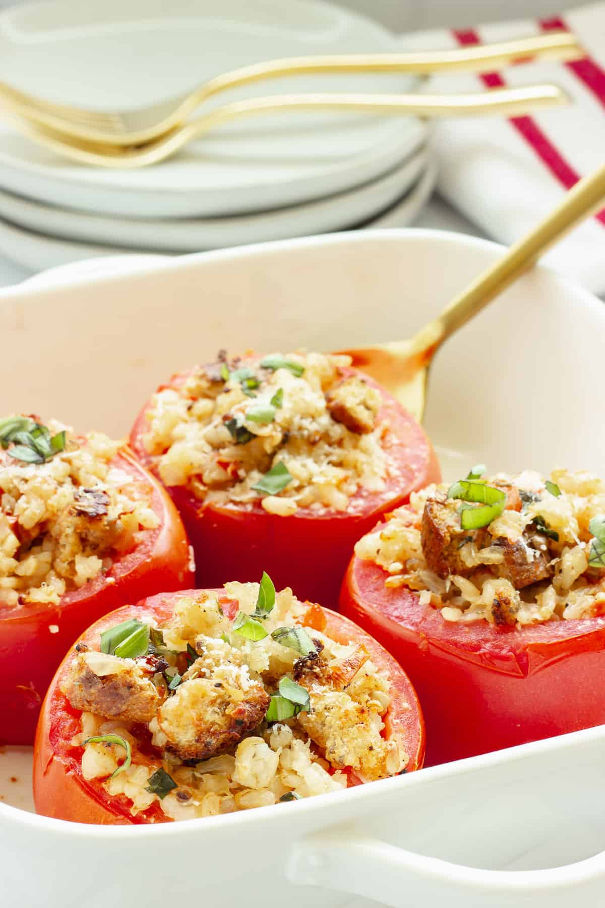 A white casserole dish with 4 baked stuffed tomatoes. A gold serving spoon, forks, and plates in the background.