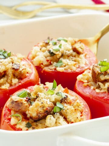 A white casserole dish with 4 baked stuffed tomatoes. A gold serving spoon and forks in the background.