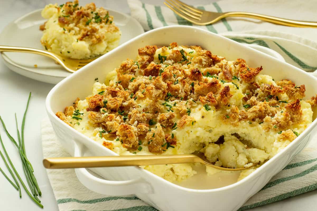 A white casserole dish with boursin mashed potatoes. A serving dish, green napkin, and bunch of chives are in the background.