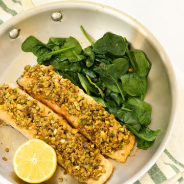 A frying pan with two pistachio crusted salmon fillets, a cut lemon, and sauteed spinach.