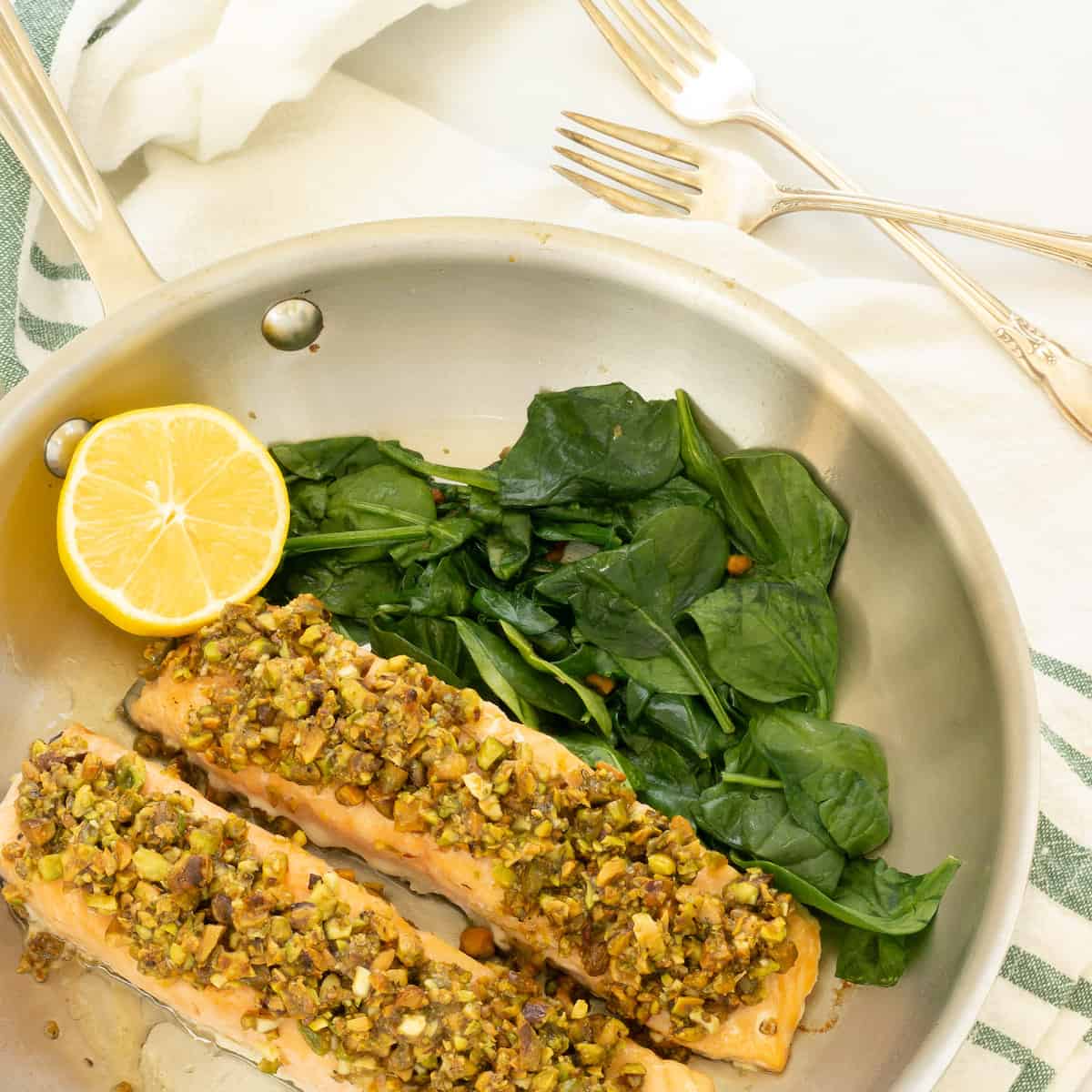 A frying pan with two pistachio crusted salmon fillets, garnished with a cut lemon, and sauteed spinach. Two forks and a green, white striped napkin are in the background.