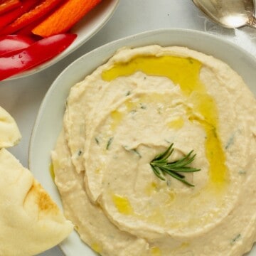 A white plate spread with white bean dip. Cut red peppers and pita bread are next to the plate.