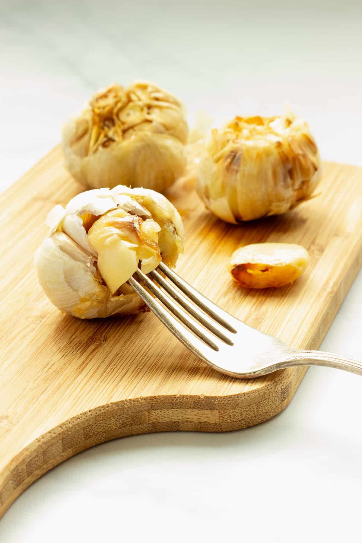 A head of air fryer roasted garlic on a cutting board. A fork is lifting a clove of garlic from the garlic head.