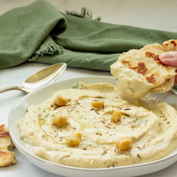 A plate of homemade hummus and a hand dipping naan bread into the hummus. A spoon and green napkin are in the background.